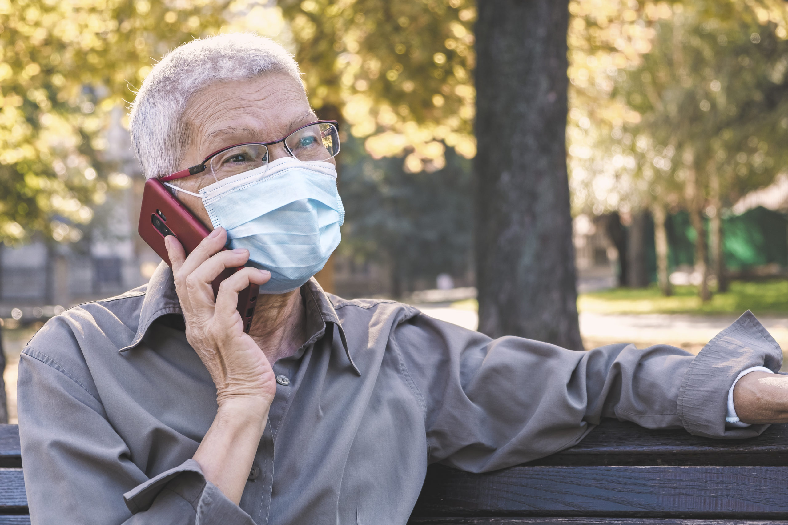 mature man with disposable mask talking on a cell phone while sitting on a park bench